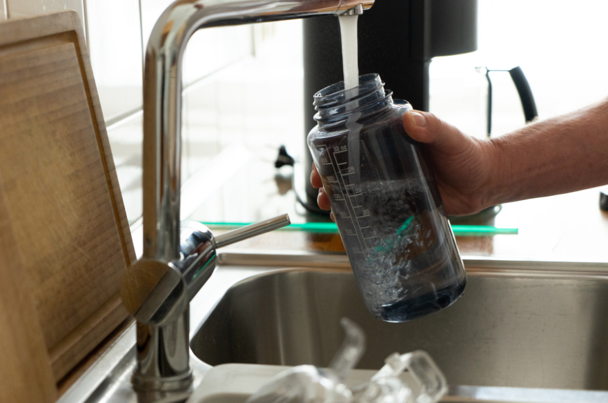 A person filling a reusable water bottle at a kitchen sink next to meal prep containers on the counter